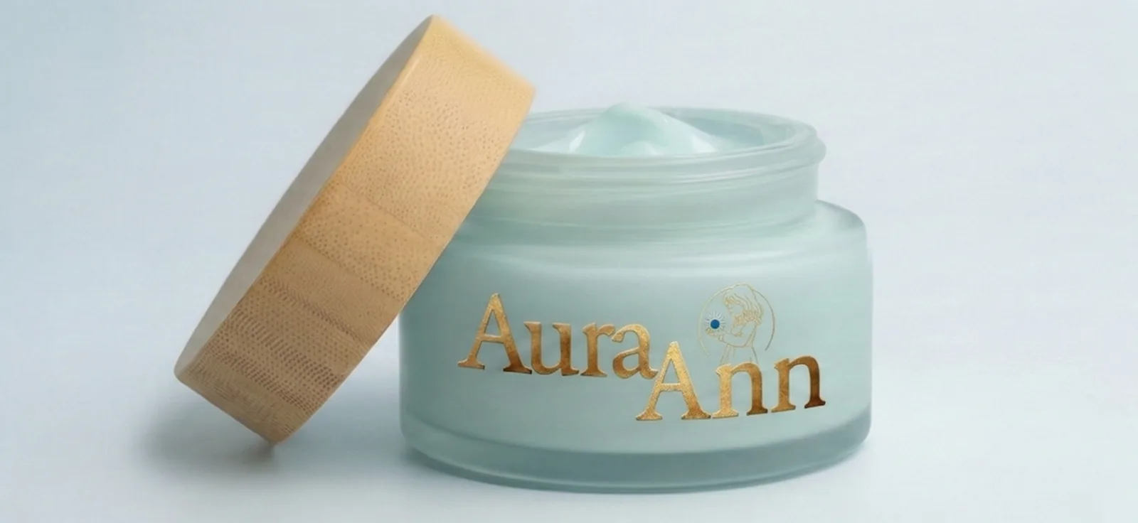 Frosted blue-green AuraAnn jar with the bamboo lid resting against it and pale blue cream visible inside, on a cool light background